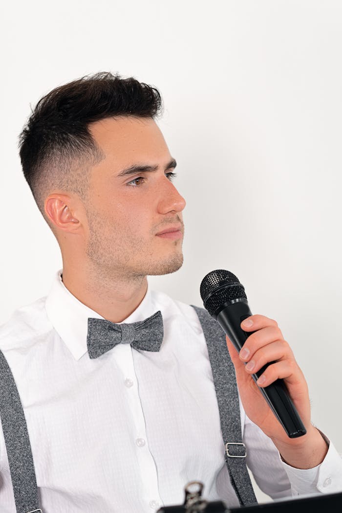 Young caucasian man in white shirt and bow tie holding a microphone, looking serious.